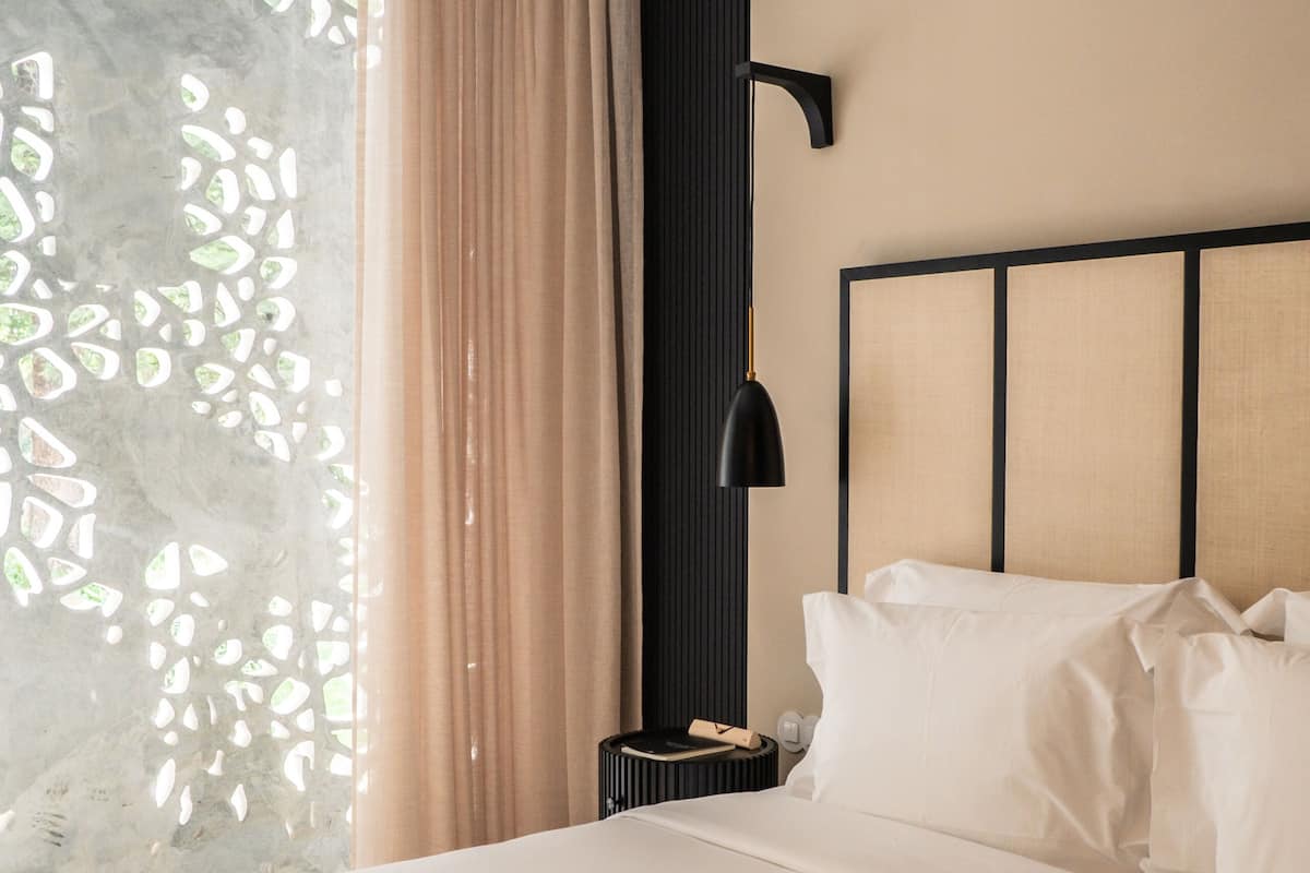 Quarto de hotel com cama, mesa de cabeceira, luminária e janela com cortinas.