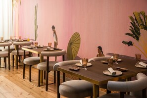 A restaurant with wooden floors, round tables with grey upholstered chairs, and a pink wall with wall art. - ARTSY Cascais (Cascais)