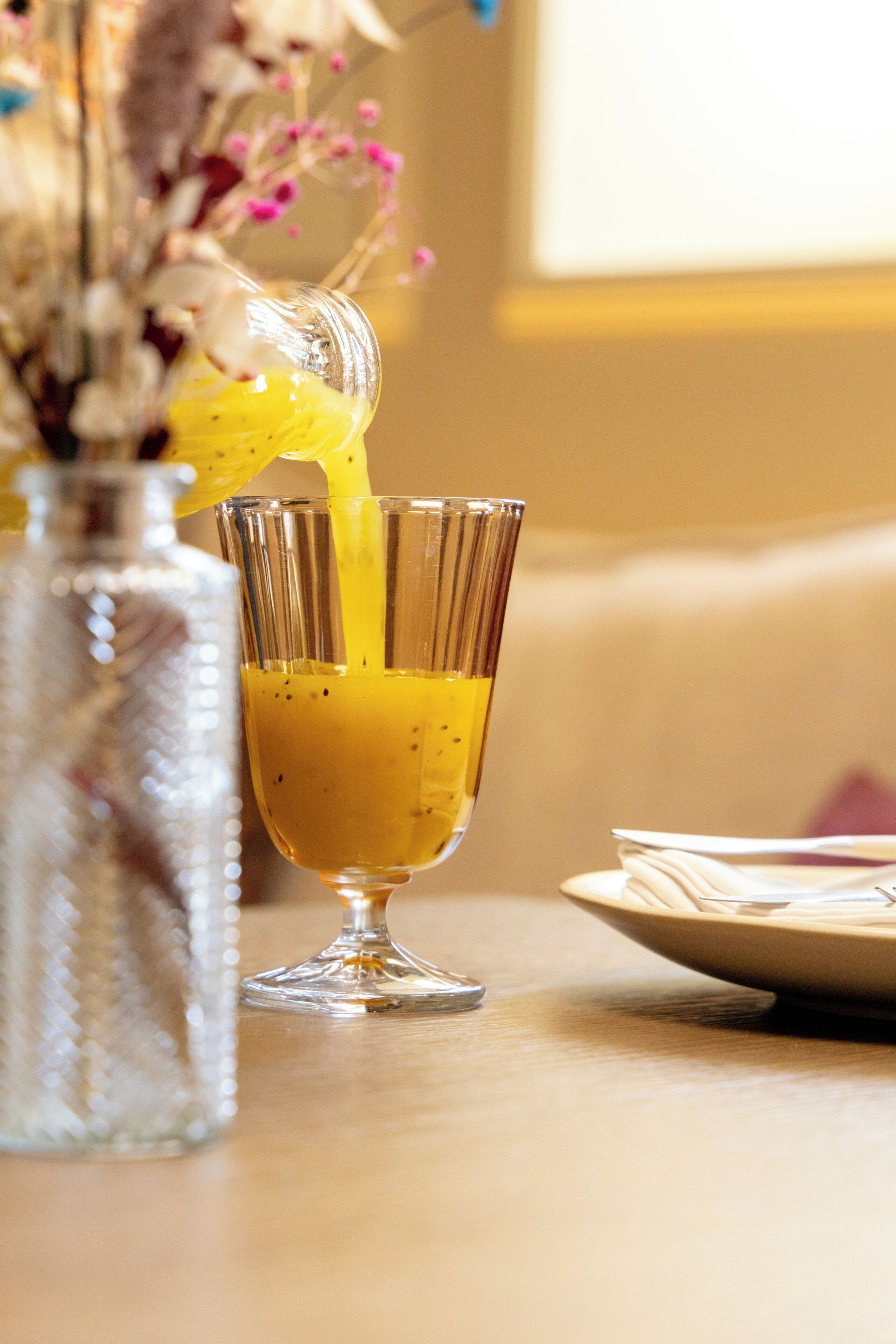 Un vaso transparente con líquido amarillo, un florero decorativo con flores secas y un plato con servilleta sobre superficie de madera.