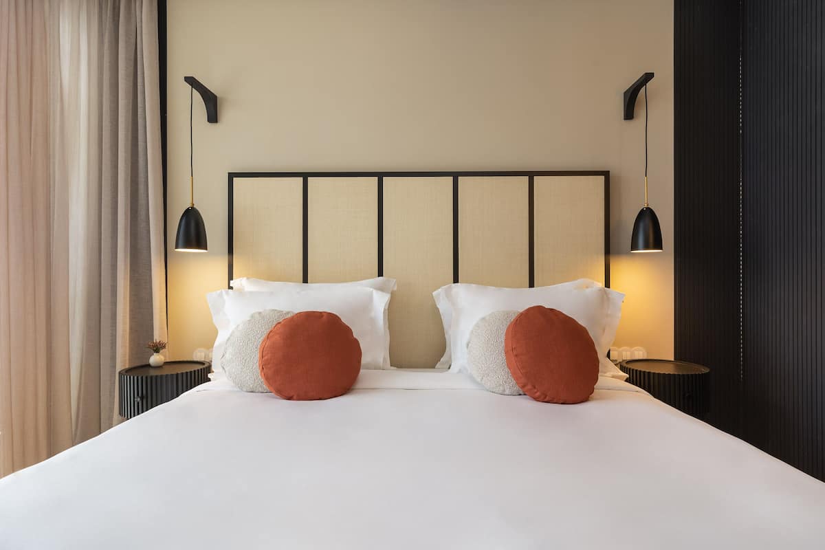 Cama bem arrumada com lençóis brancos e duas almofadas vermelhas, acompanhada por mesas de cabeceira pretas com abajures.