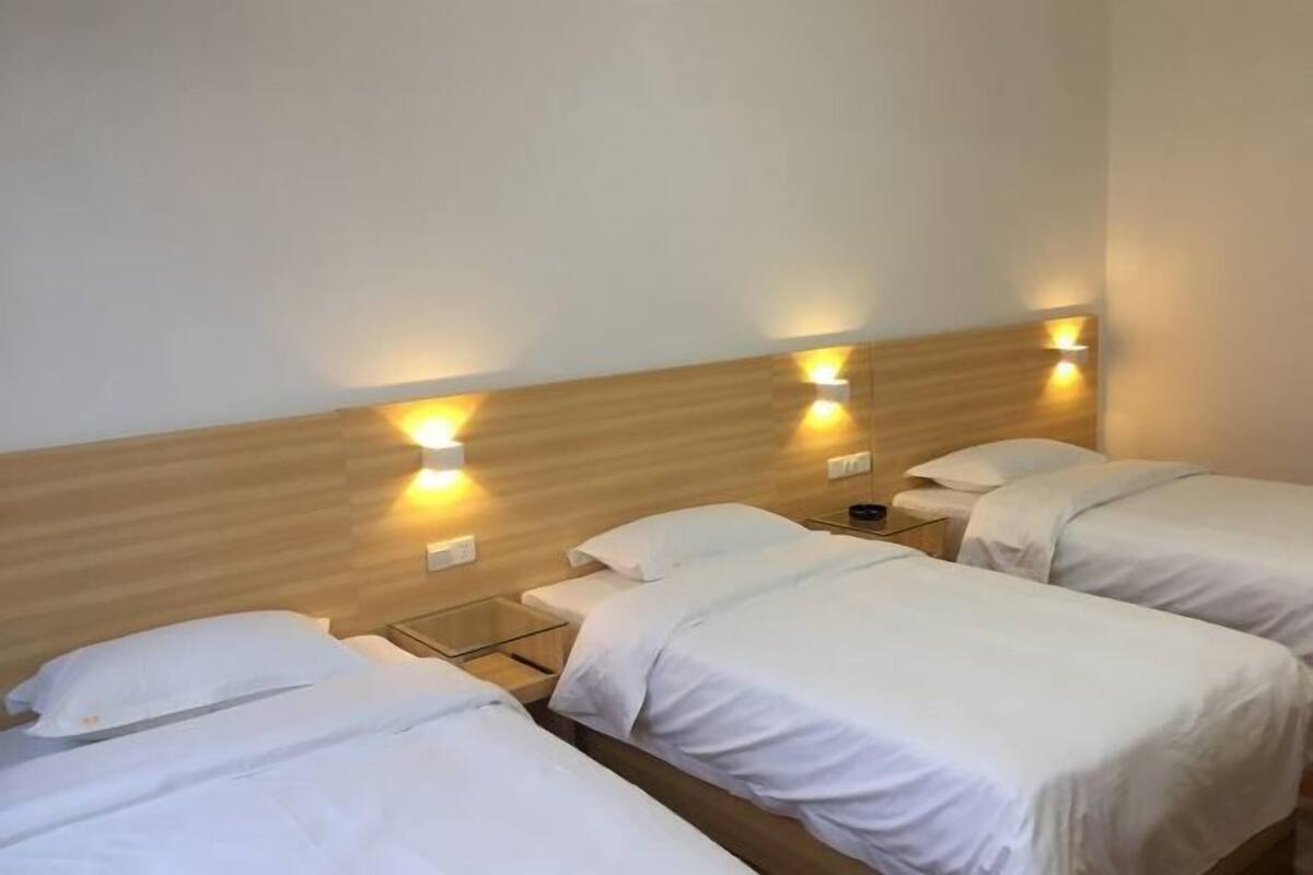 A hotel room with three single beds, wooden headboard, bedside tables, and wall-mounted lamps.