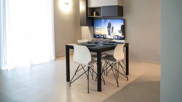 A modern dining area with a TV mounted on the wall, a dining table with chairs, and a light fixture above the table. - Dionysius Suite (Siracusa)
