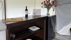 A tented area with a wooden counter, wine glasses, a bottle, a sign, and a vase with flowers.