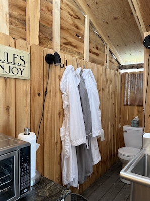 A wooden interior with a sign reading "JULES ENJOY", a microwave, a toilet, and clothes hanging on a rack.