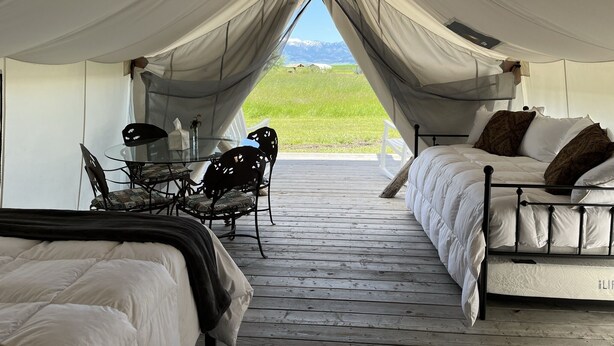 Un hébergement sous tente comprenant un lit, une table avec des chaises et une vue sur un champ verdoyant et des montagnes.