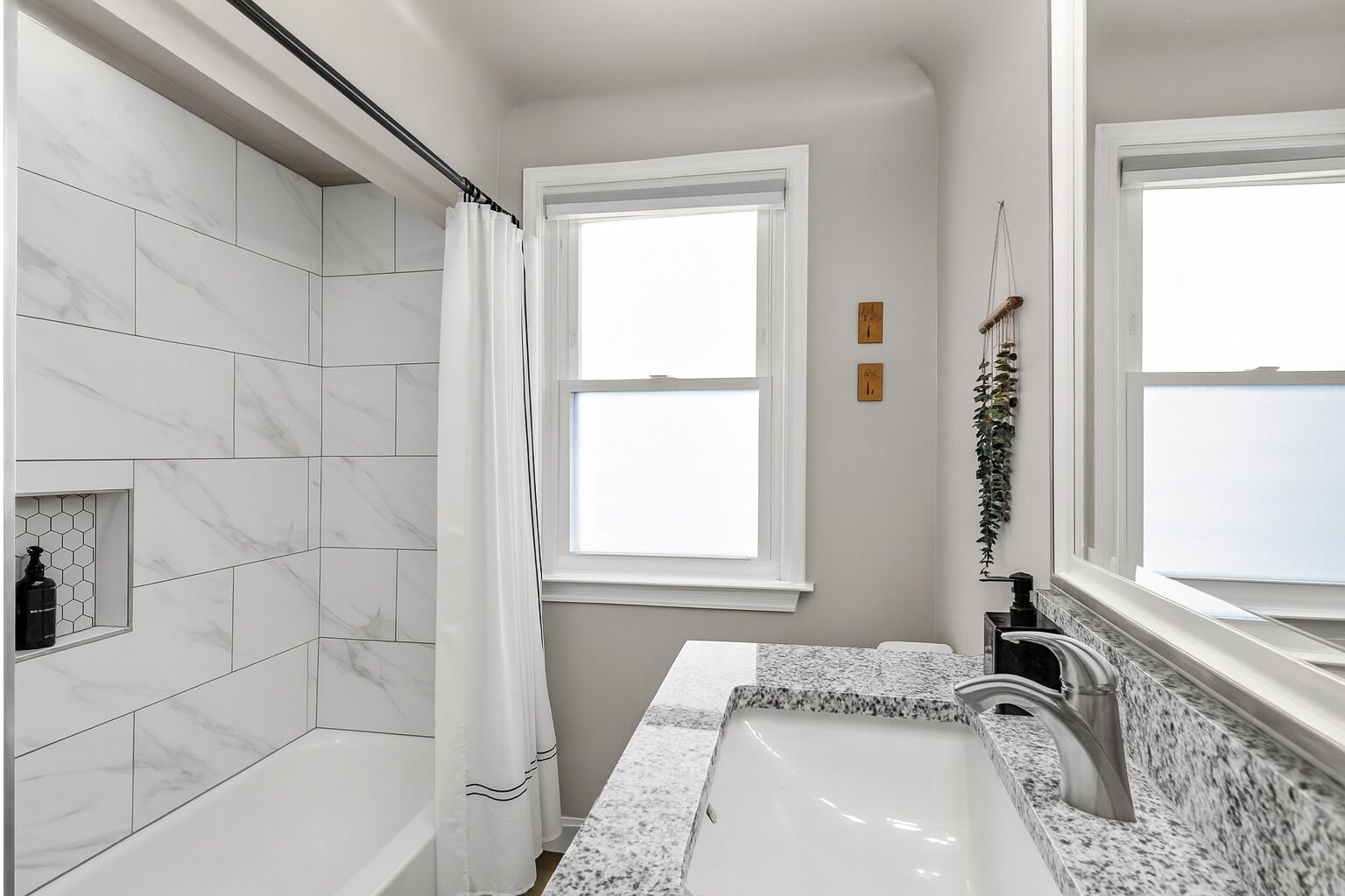 A modern bathroom with a bathtub, a sink with a granite countertop, and a window with a white curtain.