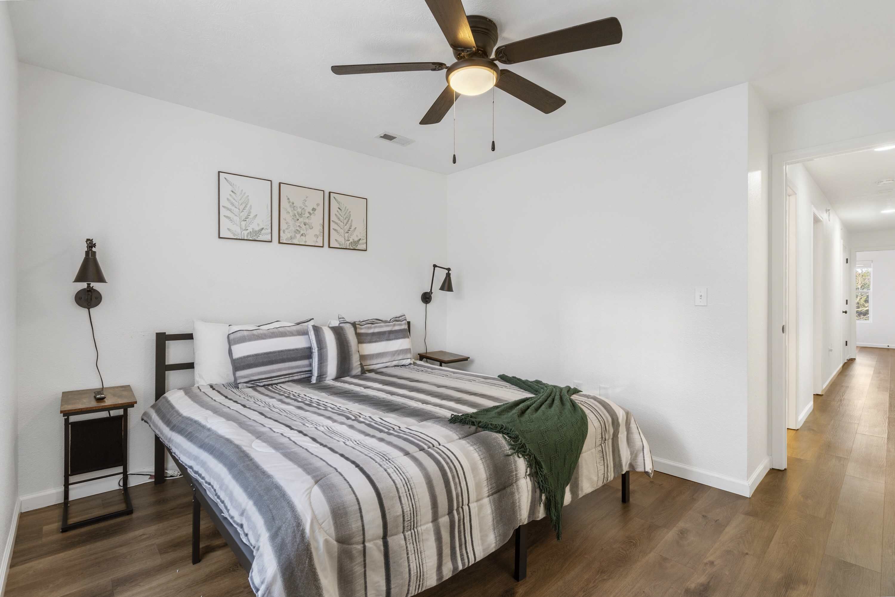 A bedroom with a bed, a ceiling fan, two bedside lamps, and framed botanical prints on the wall.