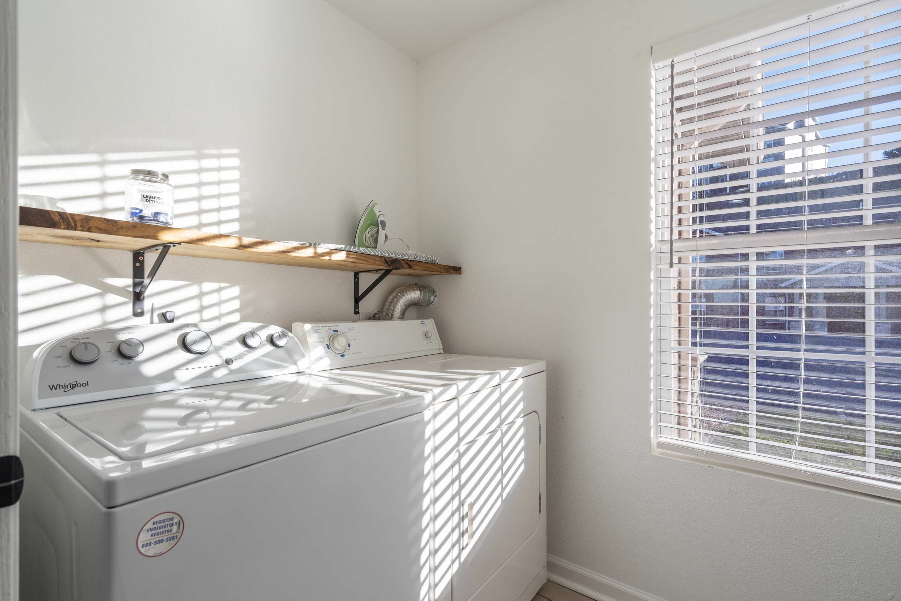 A laundry room with a front-loading washer and dryer, a shelf above the dryer, and a window with blinds.