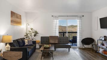 Un salón con sofá, sillón y mesa de centro, un televisor en la pared y un balcón con cortinas.