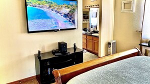 Ein Hotelzimmer mit Fernseher, der eine Strandansicht zeigt, einem Holzstuhl, einem Bett, einem kleinen schwarzen Schrank und einer Küchenzeile im Hintergrund.