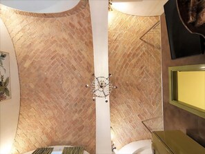Quarto com parede de tijolos, coluna branca, lustre, televisão e cama.