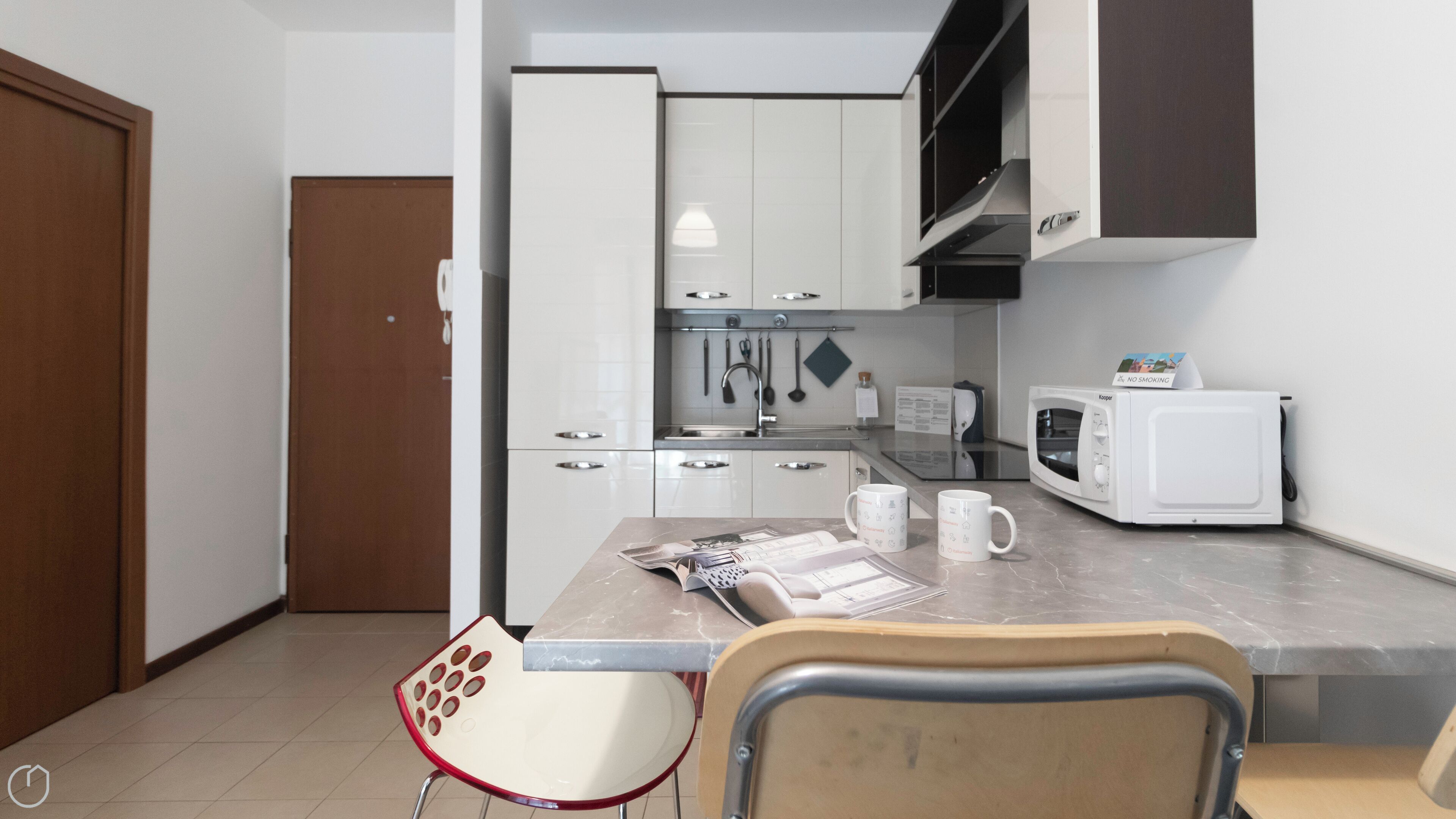 A compact kitchen with white cabinets, a microwave, and a sink. There is a chair and a table with a magazine and a mug.