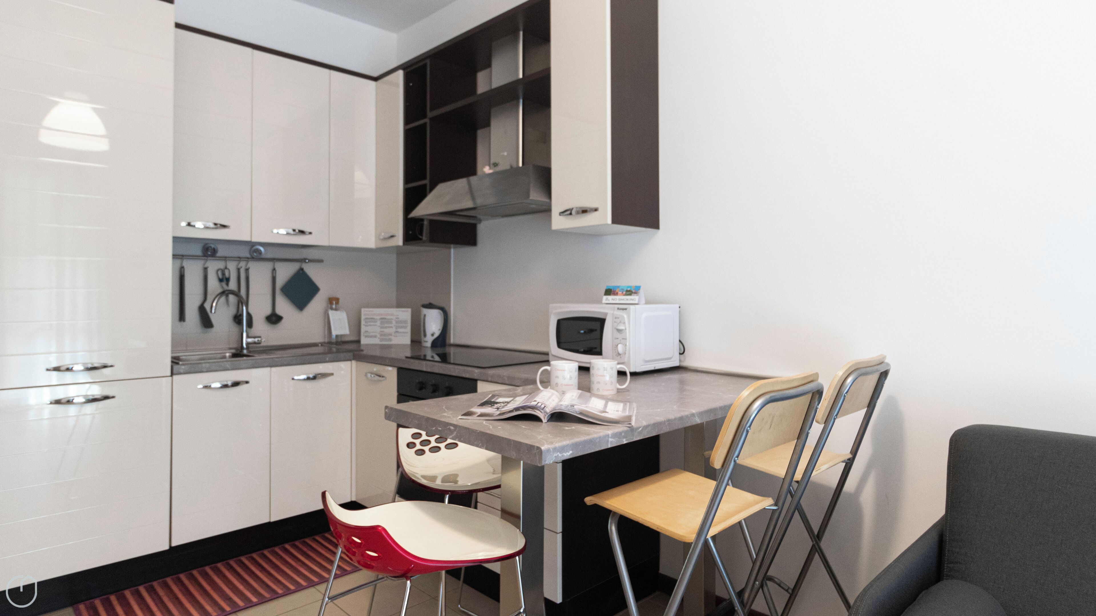 Une cuisine compacte avec des meubles blancs, un four à micro-ondes, une machine à café et une petite table à manger avec deux chaises.