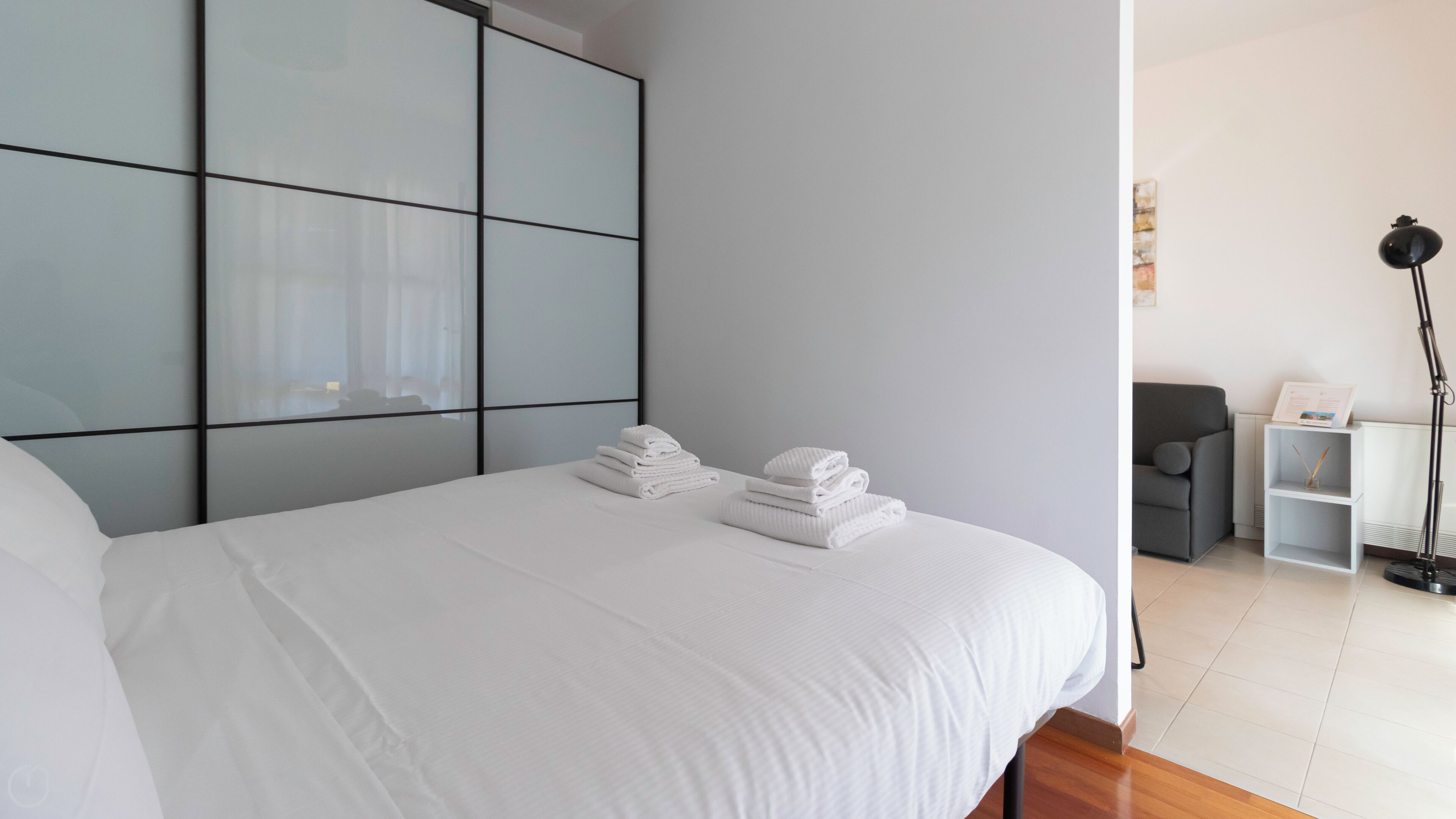 Une chambre d’hôtel moderne avec un grand lit, un canapé gris, une petite table avec une lampe et une étagère blanche.