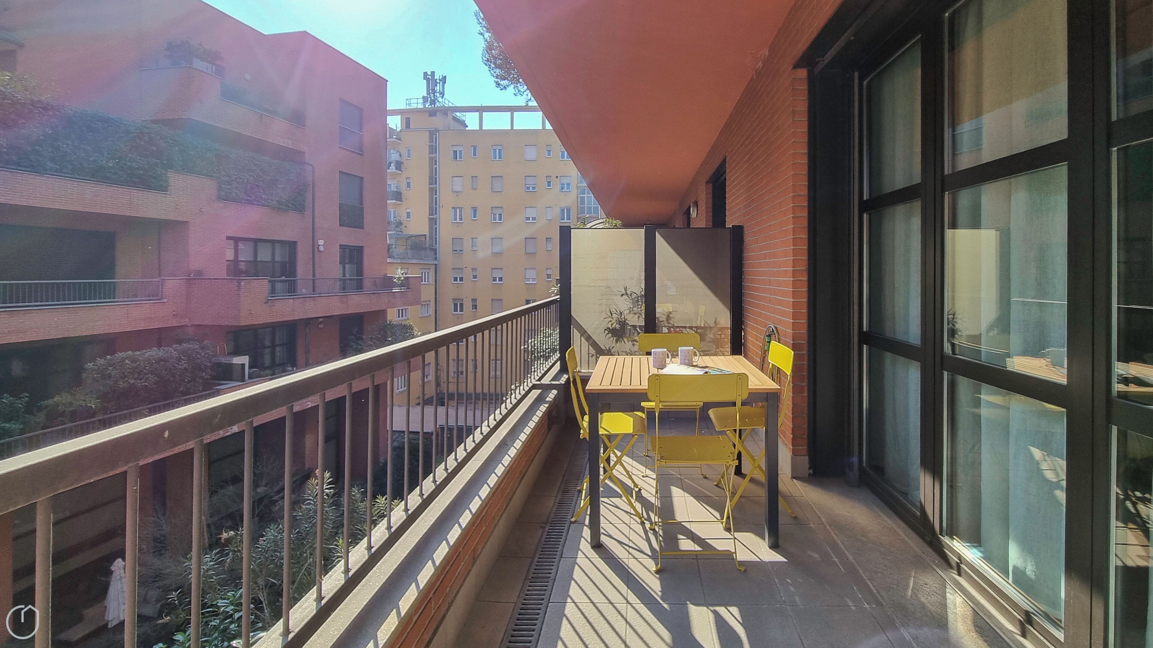 A balcony with yellow chairs and a table, overlooking a cityscape with multi-story buildings.