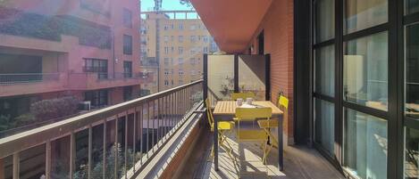A balcony with yellow chairs and a table, overlooking a cityscape with multi-story buildings.