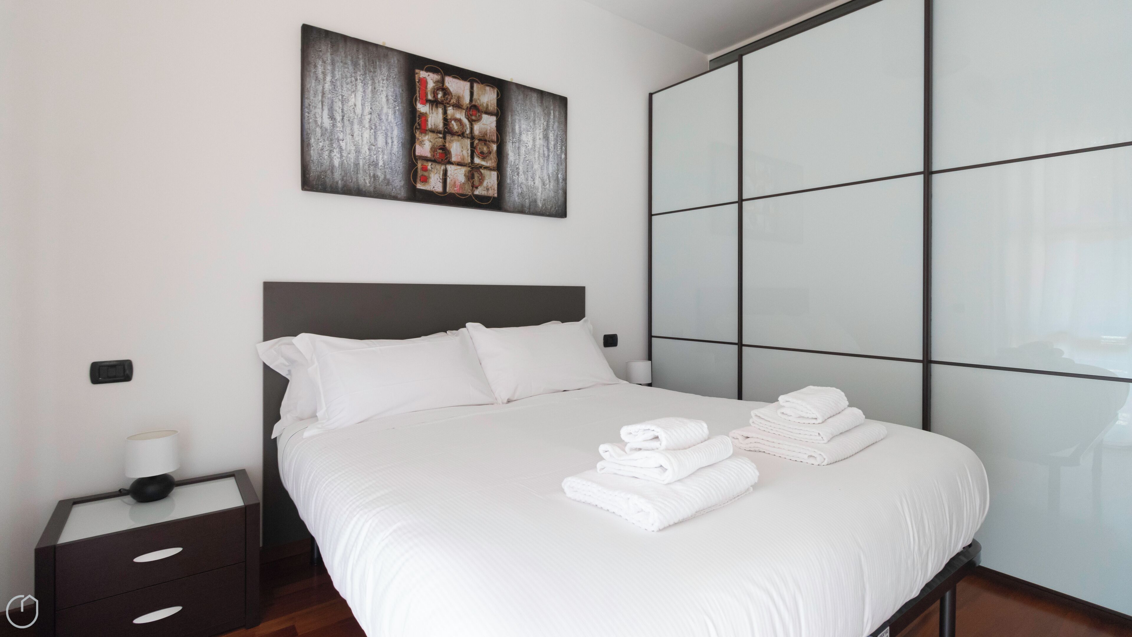 A neatly made bed with white linens and folded towels, a bedside table with a lamp, and a large abstract painting above the bed.