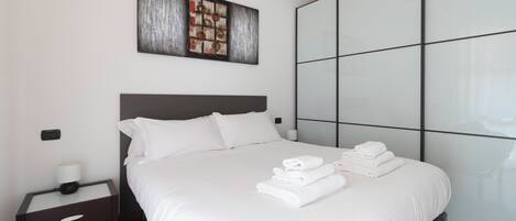A neatly made bed with white linens and folded towels, a bedside table with a lamp, and a large abstract painting above the bed.