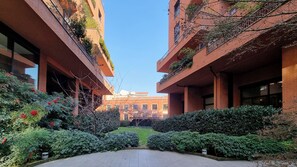 A courtyard with a central pathway flanked by brick buildings with balconies, surrounded by well-maintained hedges and flowering plants.