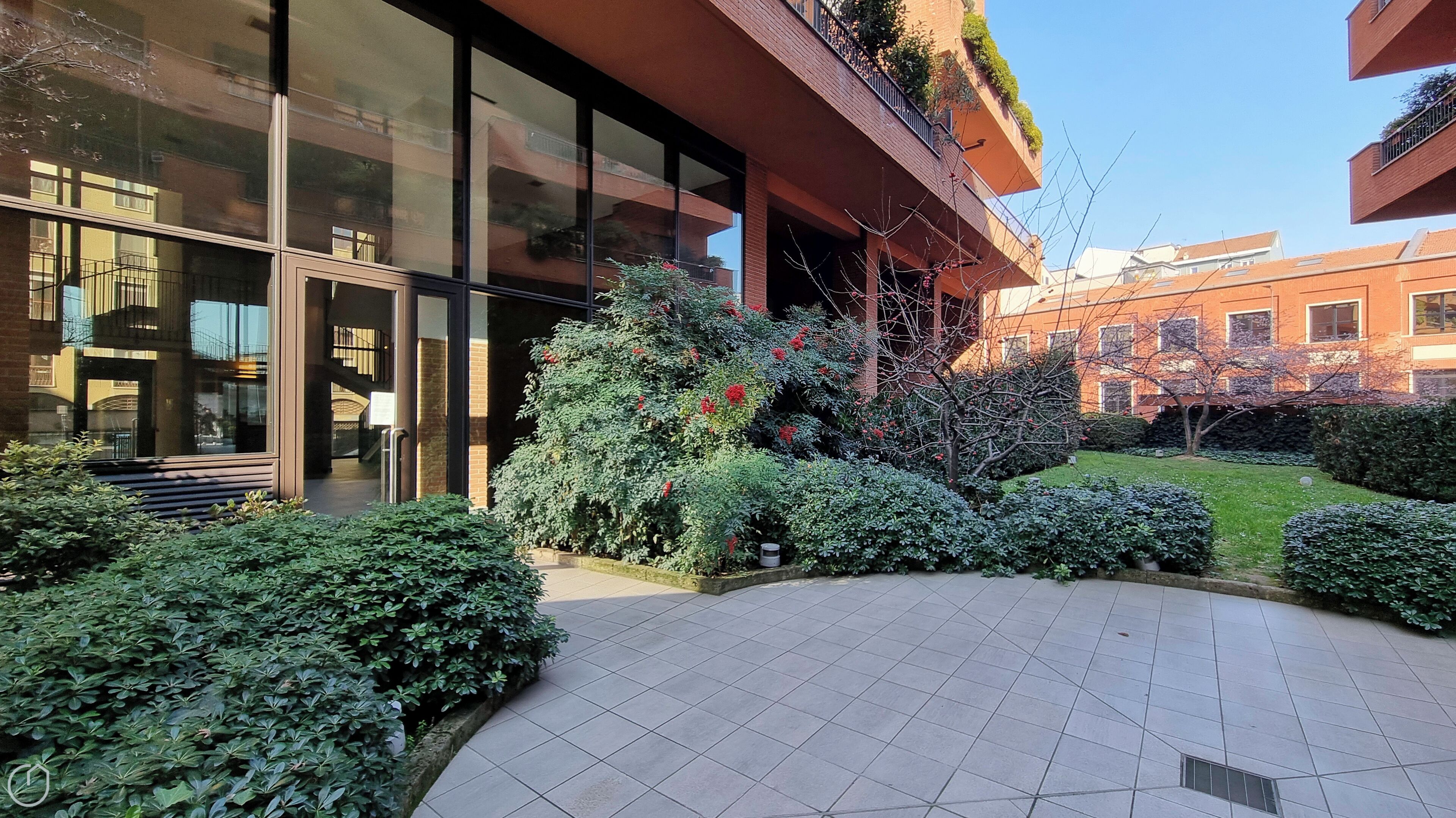 A modern building with large glass windows, a tiled courtyard, and well-maintained greenery.
