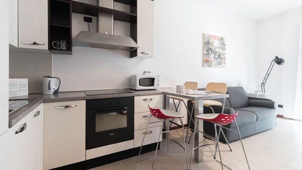 A modern kitchen with a stove, microwave, and kettle. There are two red chairs and a grey sofa in the living area.