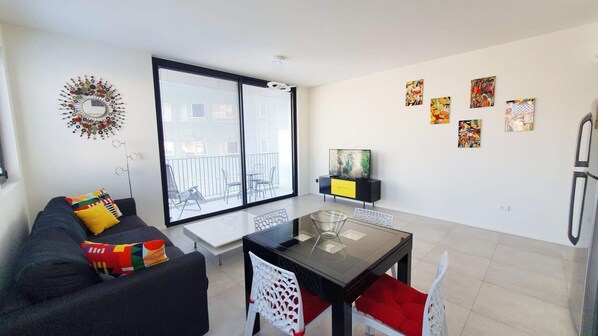 Een moderne woonkamer met een zwarte bank, kleurrijke kussens, een glazen salontafel en een eethoek met een witte stoel en een rode kussentje.