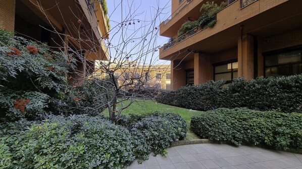 A courtyard with a tiled walkway, surrounded by dense shrubbery and a small tree with red berries. - Italianway Easy - Zuretti 32 (Milan)