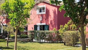 Un bâtiment rouge avec une fenêtre blanche et des volets verts, entouré d’arbres et d’une pelouse bien entretenue.
