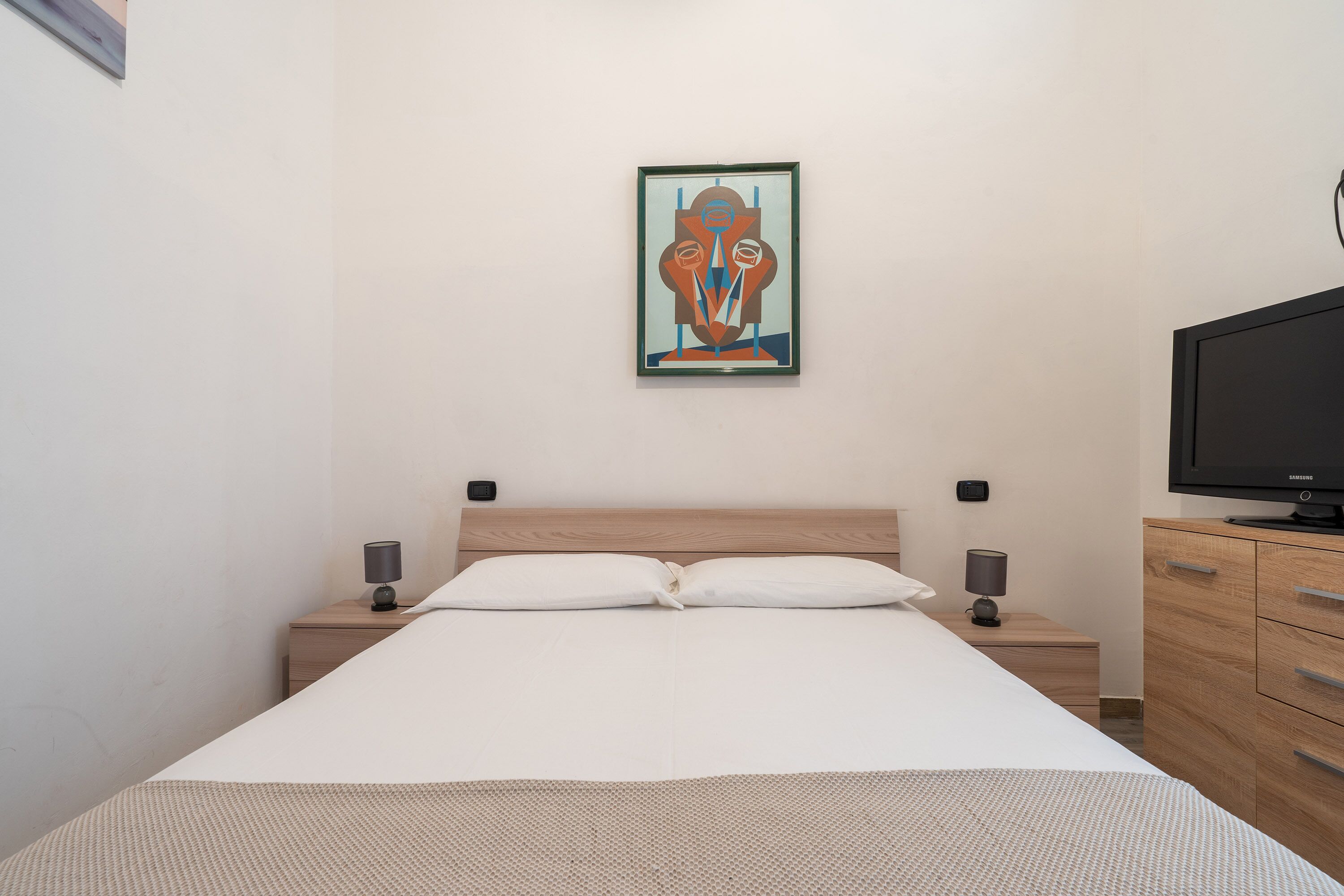 Ein ordentlich bezogenes Bett mit weißer Bettwäsche, einem Holzkopfstück und einem einzelnen Kunstwerk an der Wand.