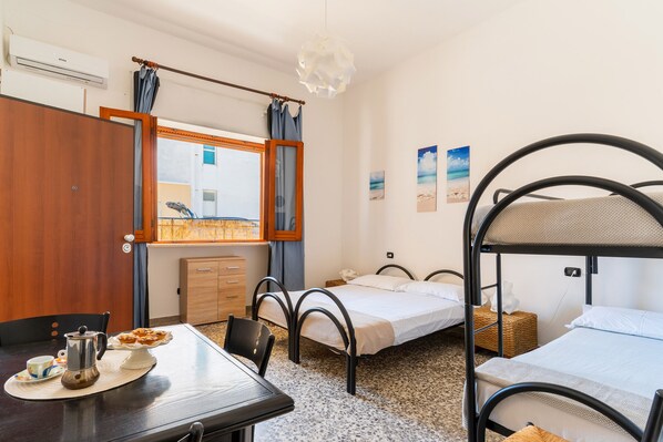 A room with bunk beds, a dining table with a coffee pot and pastries, a wooden cabinet, and a window with curtains.