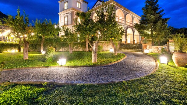 A well-lit, historic building with a cobblestone pathway leading to it, surrounded by lush greenery and trees. - Villa Artemisia in Piano di Sorrento (Piano di Sorrento)
