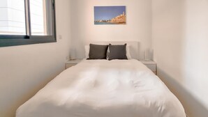 Una cama bien hecha con ropa de cama blanca y dos almohadas oscuras, una mesita de noche con una lámpara, y un cuadro en la pared con la imagen de un edificio costero.