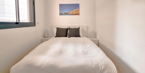 A neatly made bed with white bedding and two dark pillows, a small bedside table with a lamp, and a framed picture of a coastal building on the wall. - Ben Yehuda Nuage in Tel Aviv-yafo (Tel Aviv)