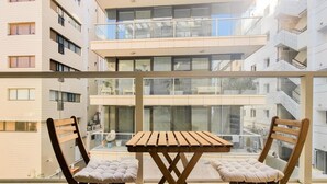Un balcón con una mesa de madera y dos sillas con cojines, con vistas a un edificio residencial de varios pisos con balcones y ventanas.
