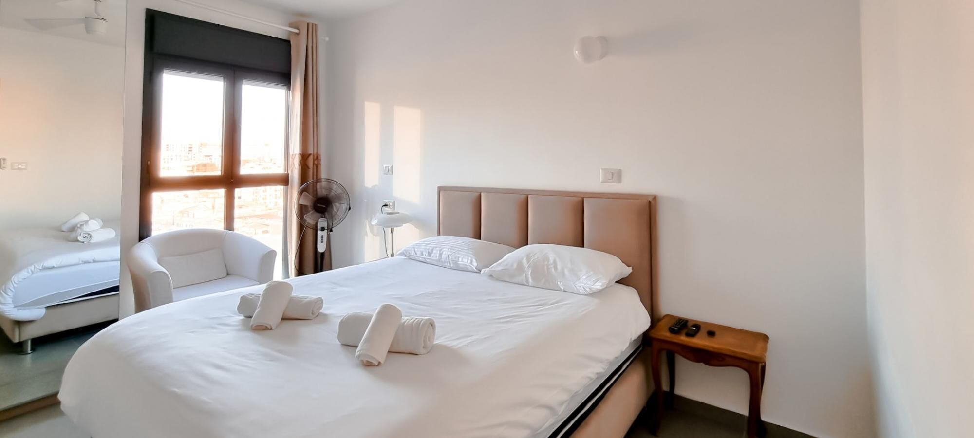 A neatly made bed with white linens and towels, a wooden nightstand with a remote control, a white armchair, and a window with curtains.