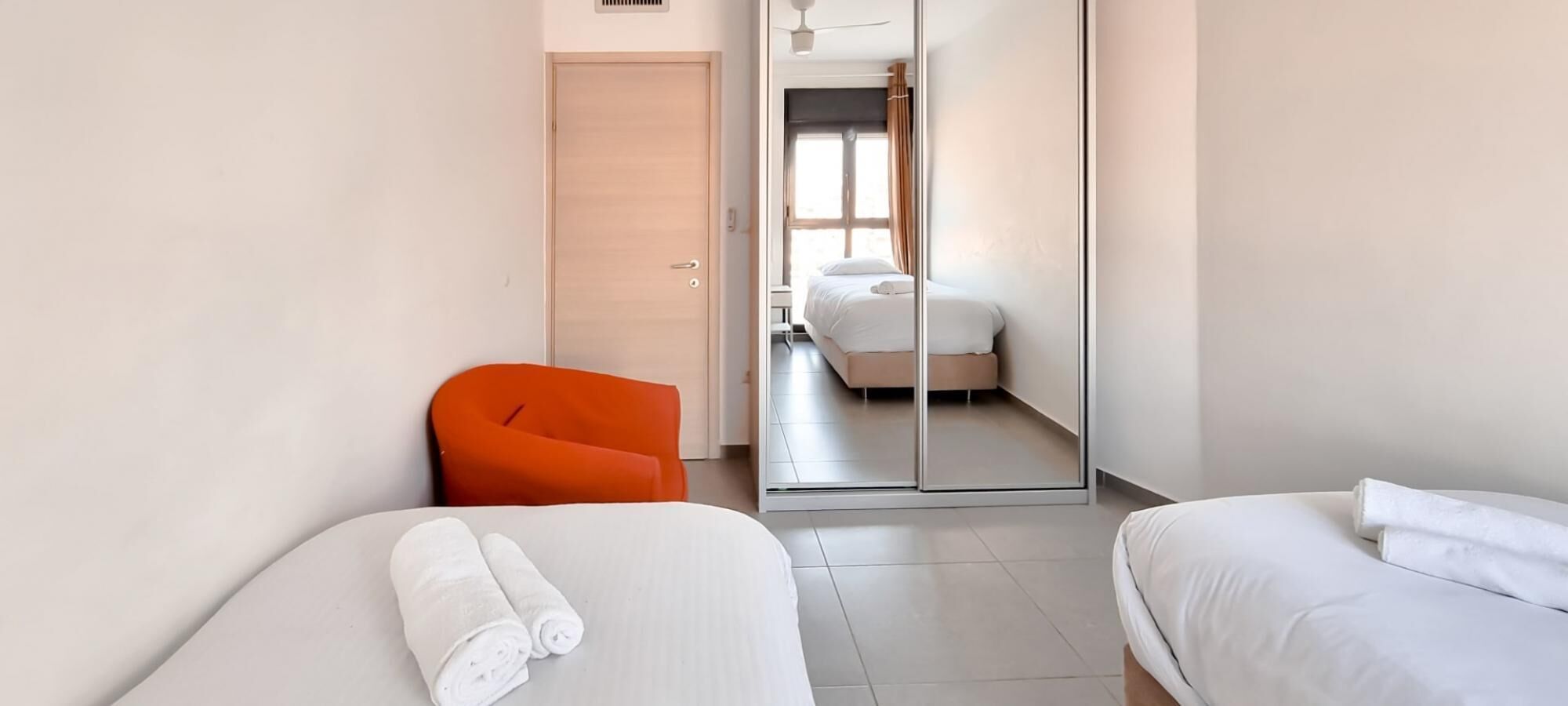 A hotel room with two beds, a sliding door leading to a bathroom, and a bright orange chair.