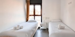 A hotel room with two single beds, a small bedside table, a lamp, and a window with curtains.