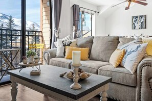 A cozy living room with a grey sofa, a white table, and a view of snow-covered mountains through large windows. - Cozy PC Condo Top Floor Studio (Park City)