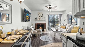 A modern living room with a fireplace, a dining area with a long table set for four, and a cozy seating area with a sofa and ottoman.