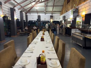 Restaurante com mesas longas e compartilhadas para refeições, espaço para buffet e balcão de atendimento.