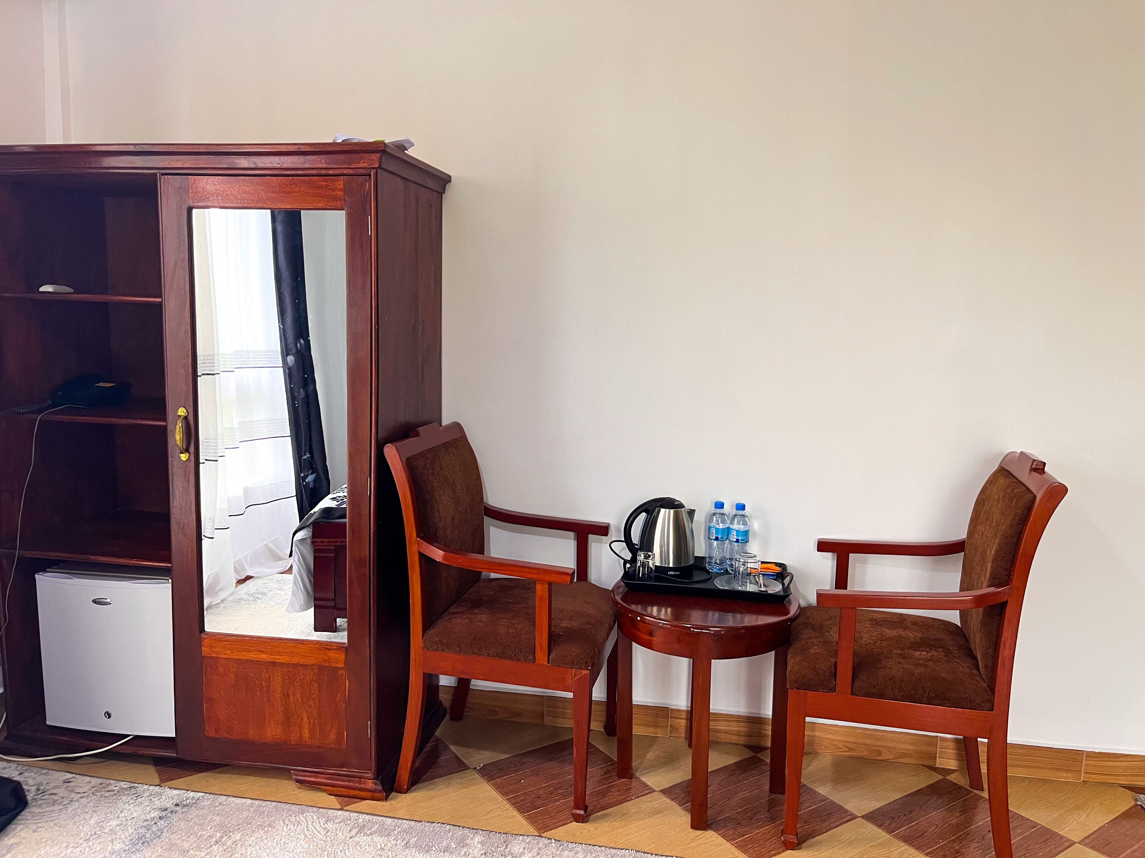 A room with a wooden cabinet, two brown upholstered chairs, a small round table with a kettle, water bottles, and glasses.