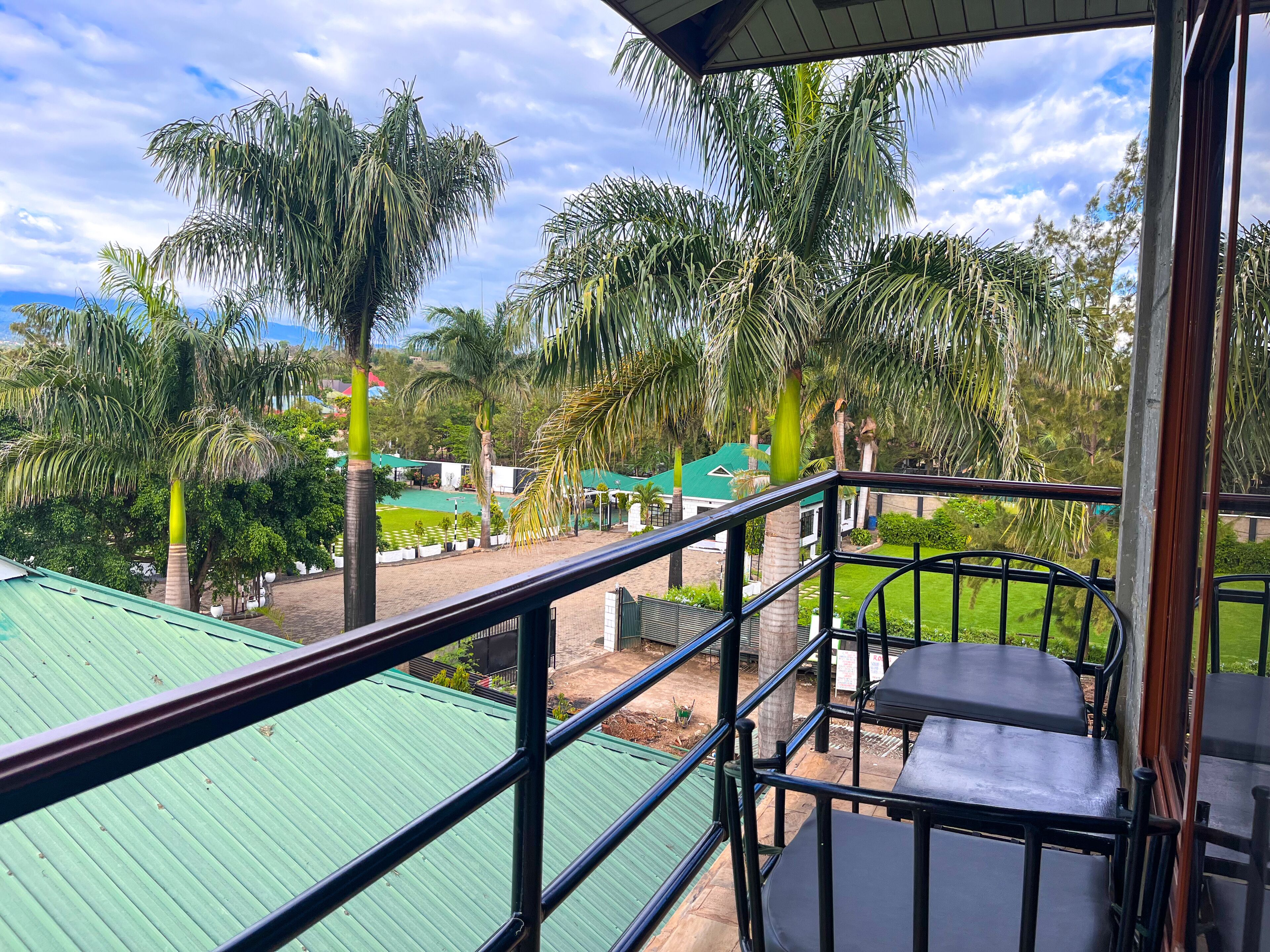 A balcony with a view of a green-roofed building, palm trees, and a grassy area.