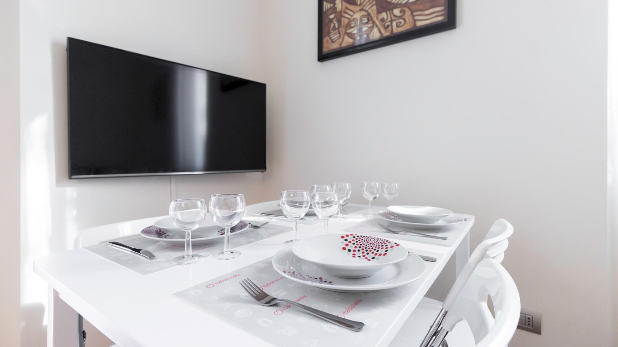 Een eettafel gedekt met witte borden, glazen en bestek, met een tv aan de muur erboven.