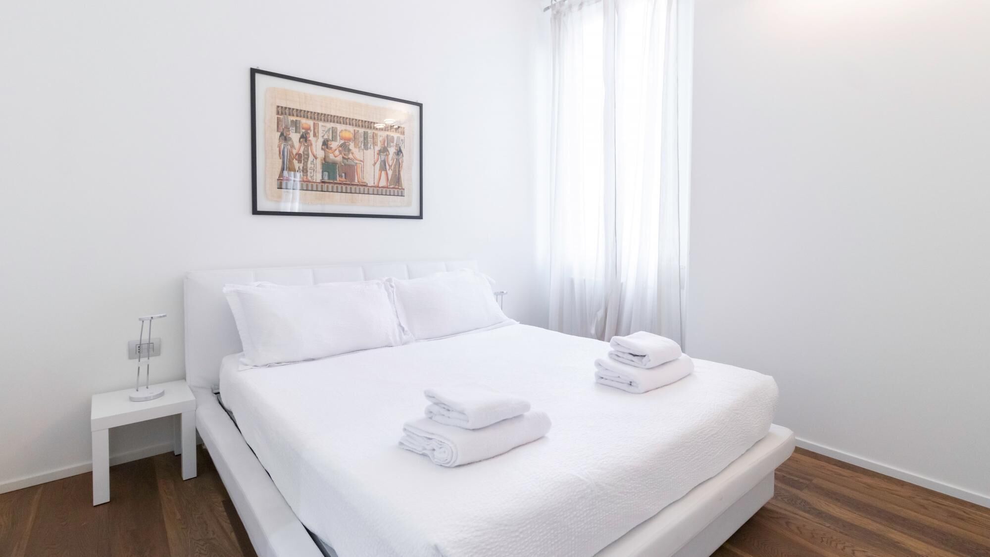 A neatly made bed with white linens and towels, a small bedside table with a glass of water, and a framed picture of ancient Egyptian figures on the wall.
