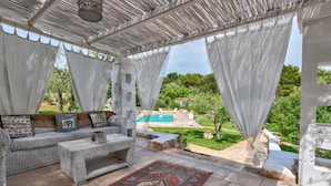 A covered outdoor seating area with a pool view, stone flooring, and a woven sofa with cushions. - Pigreco 6 2 (Ostuni)