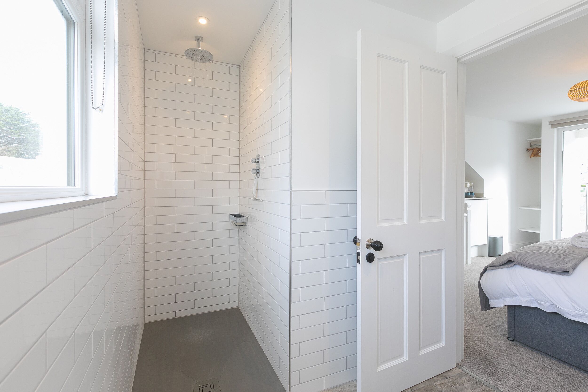 Um banheiro moderno com paredes revestidas de azulejos brancos, chuveiro e porta que dá para um quarto com cama e mesa de cabeceira.