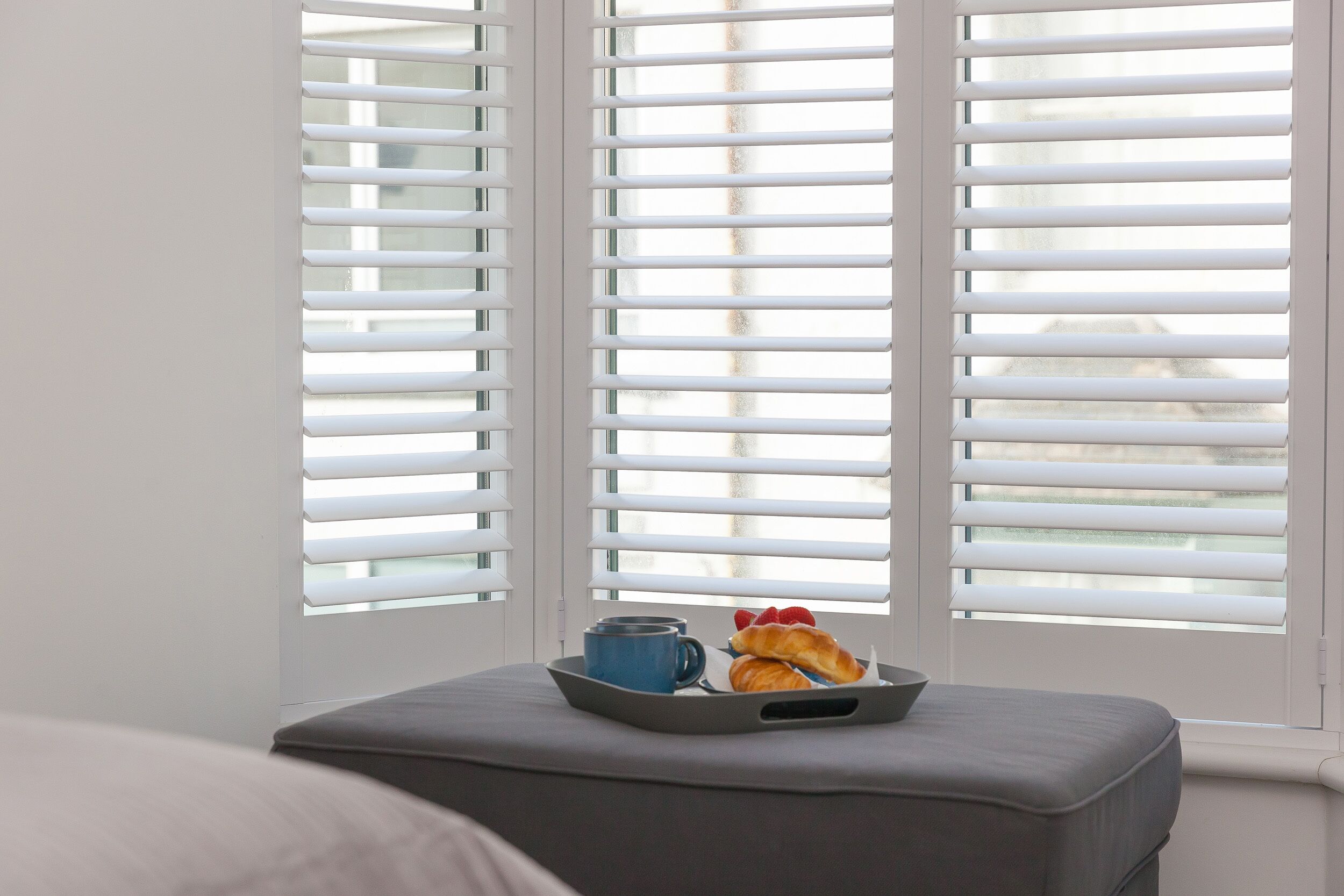 A window with white blinds, a tray with croissants and a blue mug on a grey ottoman.