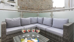A corner seating area with a cushioned sofa, a glass coffee table with snacks and drinks, and a stone building in the background. - Pebbles (St Ives)