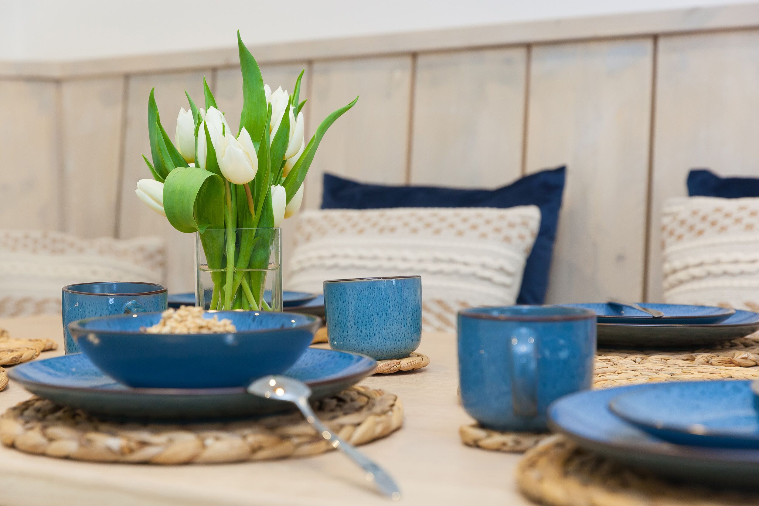 Una mesa de comedor dispuesta con platos, cuencos y tazas de cerámica azul, junto a una jarra de vidrio con tulipanes blancos.