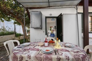 Uma área de refeições ao ar livre com uma mesa posta para dois, coberta por um toldo, e uma cadeira à esquerda.
